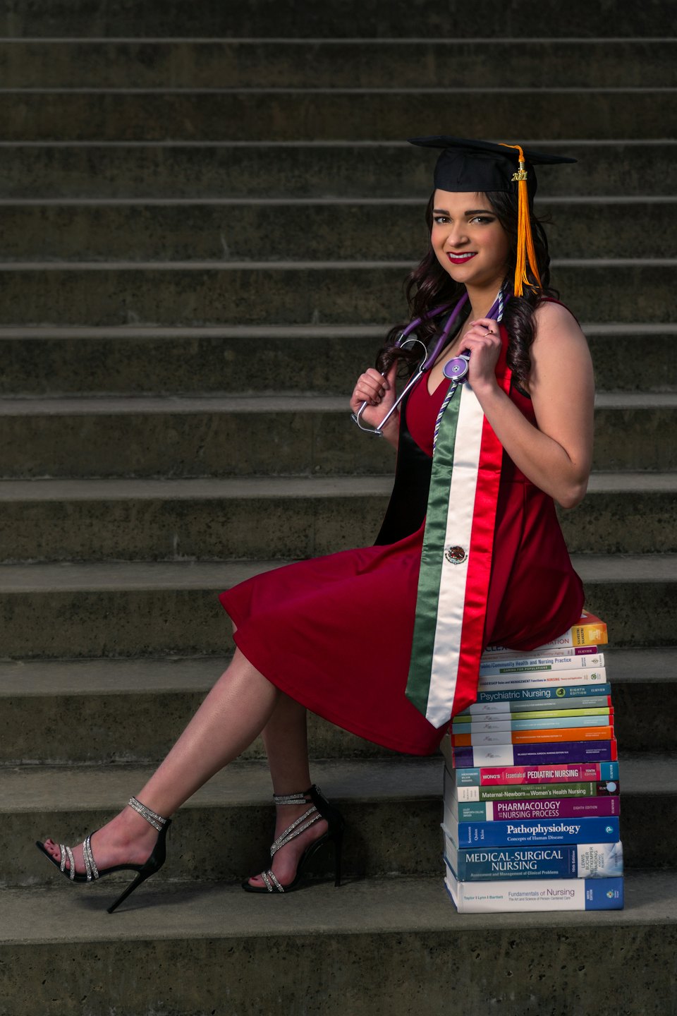 Woman in graduation cap and gown with stethoscope smiles on nursing books.